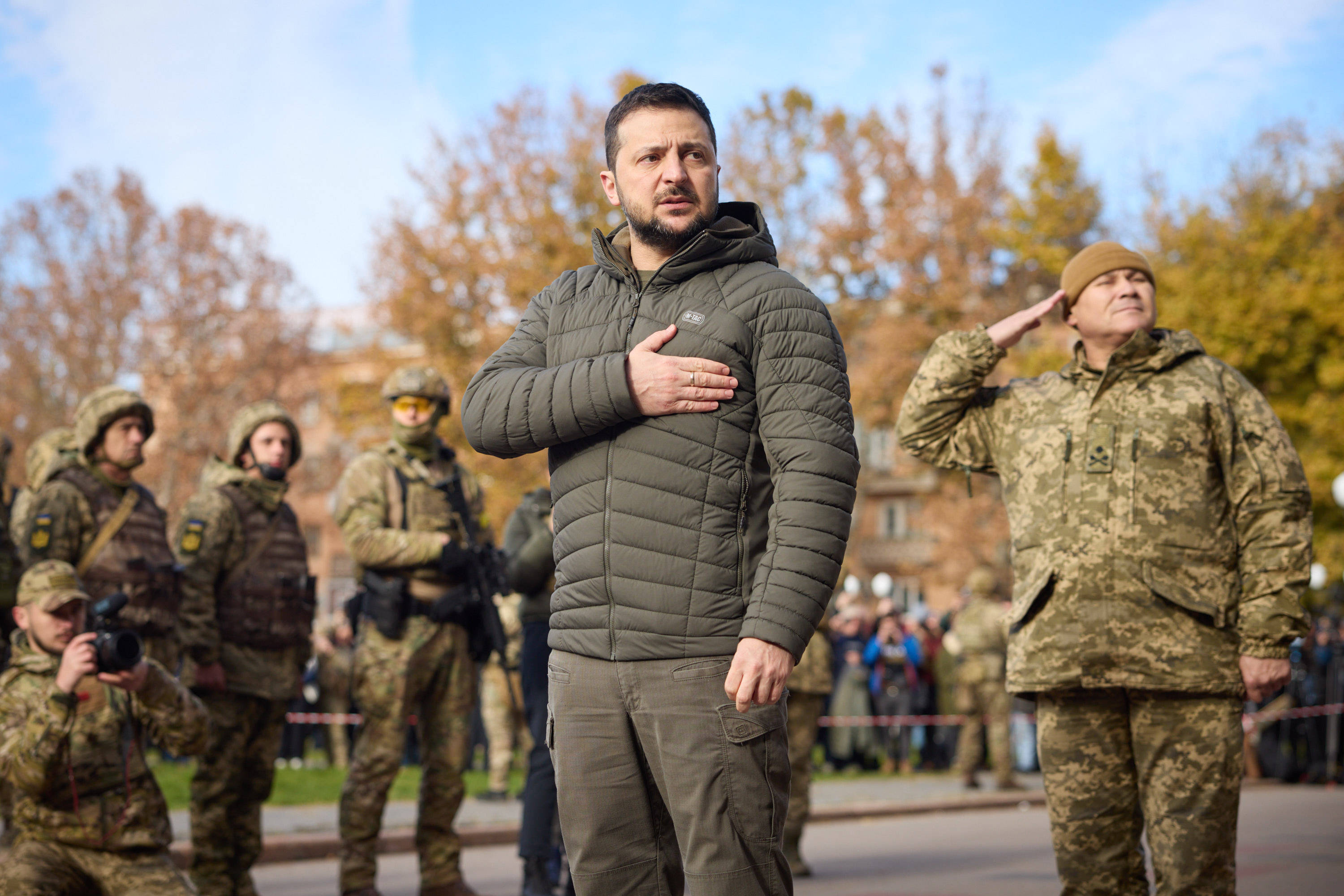 An image of the Ukrainian president with his hand over his heart standing in formation with soldiers